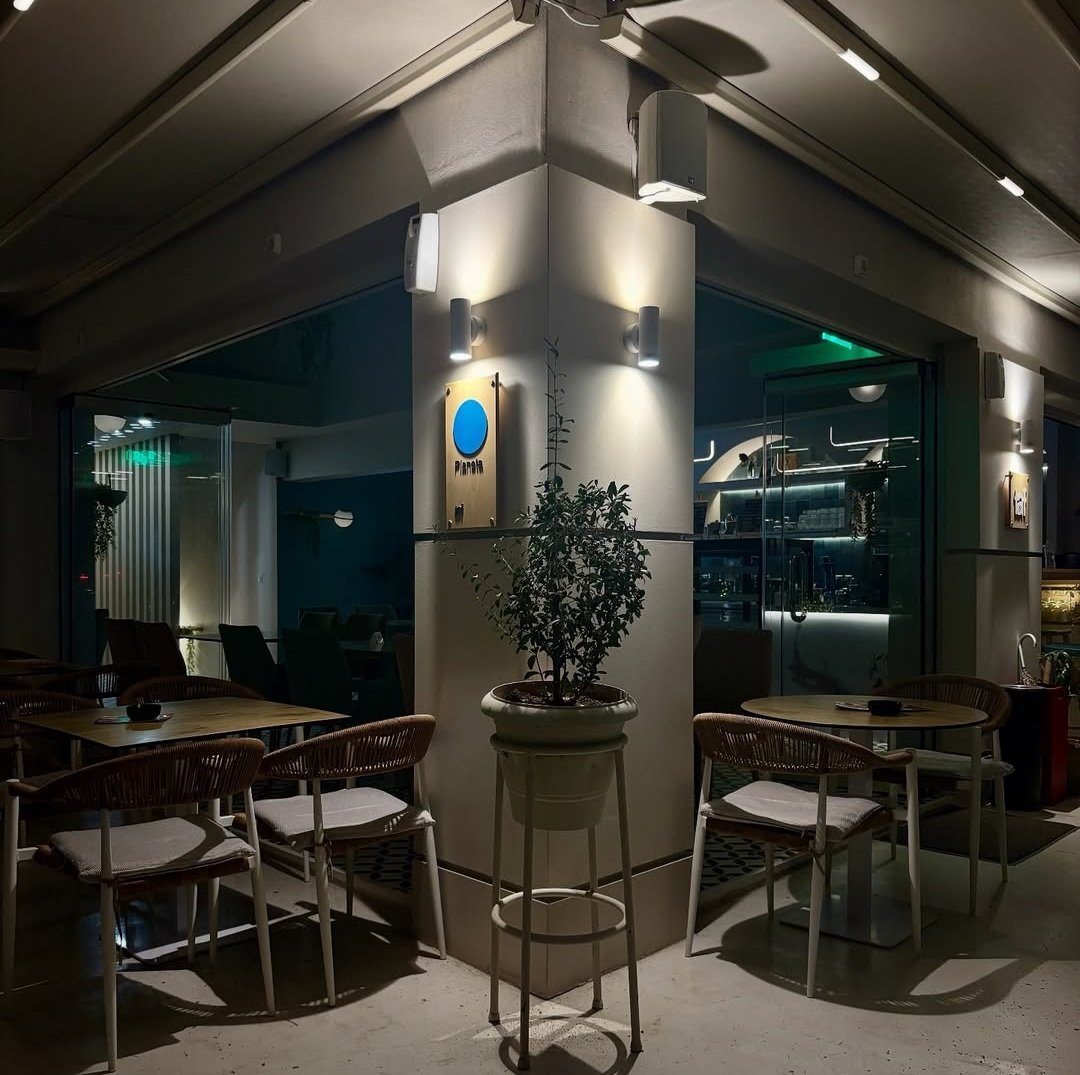 Modern office break room with teal accent walls, wooden desks and chairs, central support column with potted plant, and ambient overhead lighting