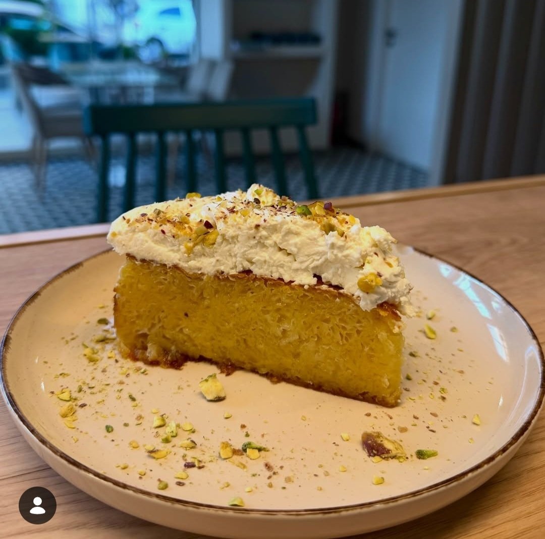 A slice of golden yellow cake with white frosting and pistachio crumble topping on a beige plate, served in a modern dining room